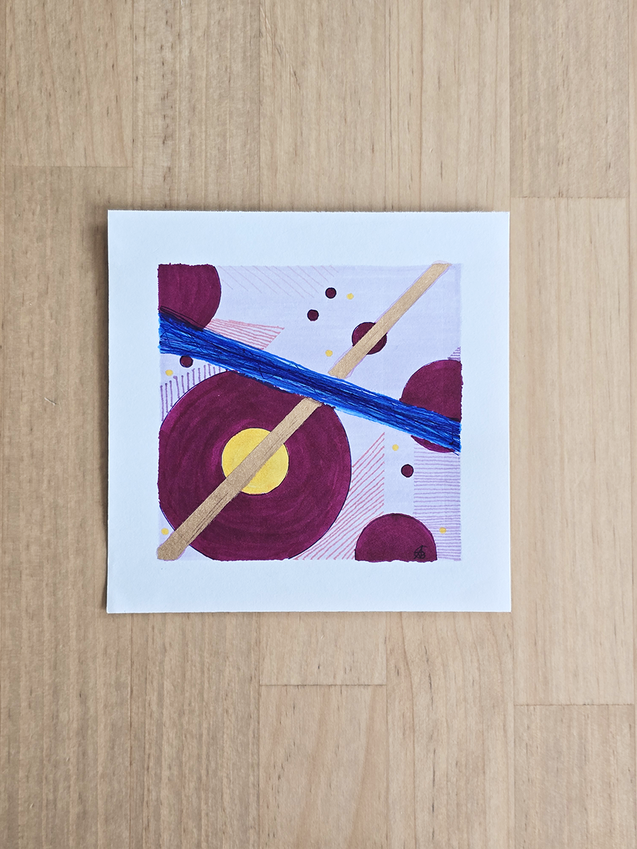 An original abstract drawing featuring geometric shapes and brushstrokes in purple, blue, yellow, and copper on a white paper border, displayed flat on a light wood countertop.