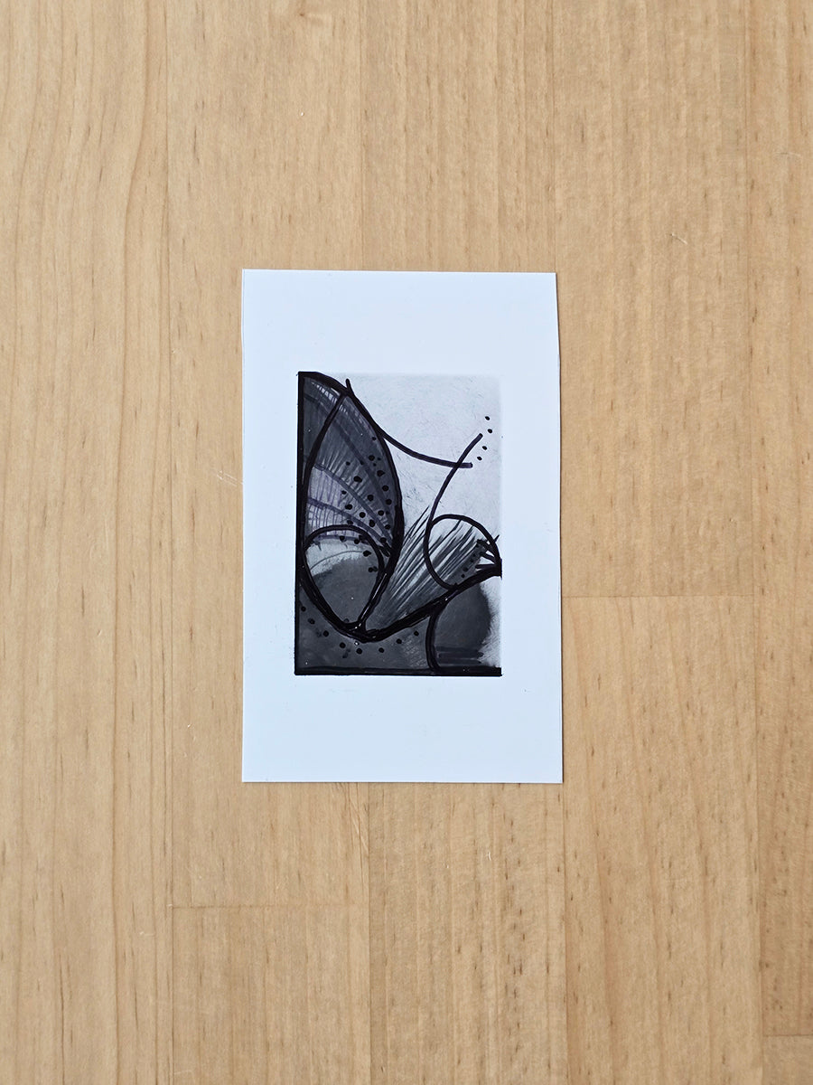 A small abstract black, gray, and white original drawing on a white paper card, placed on a light wood surface.